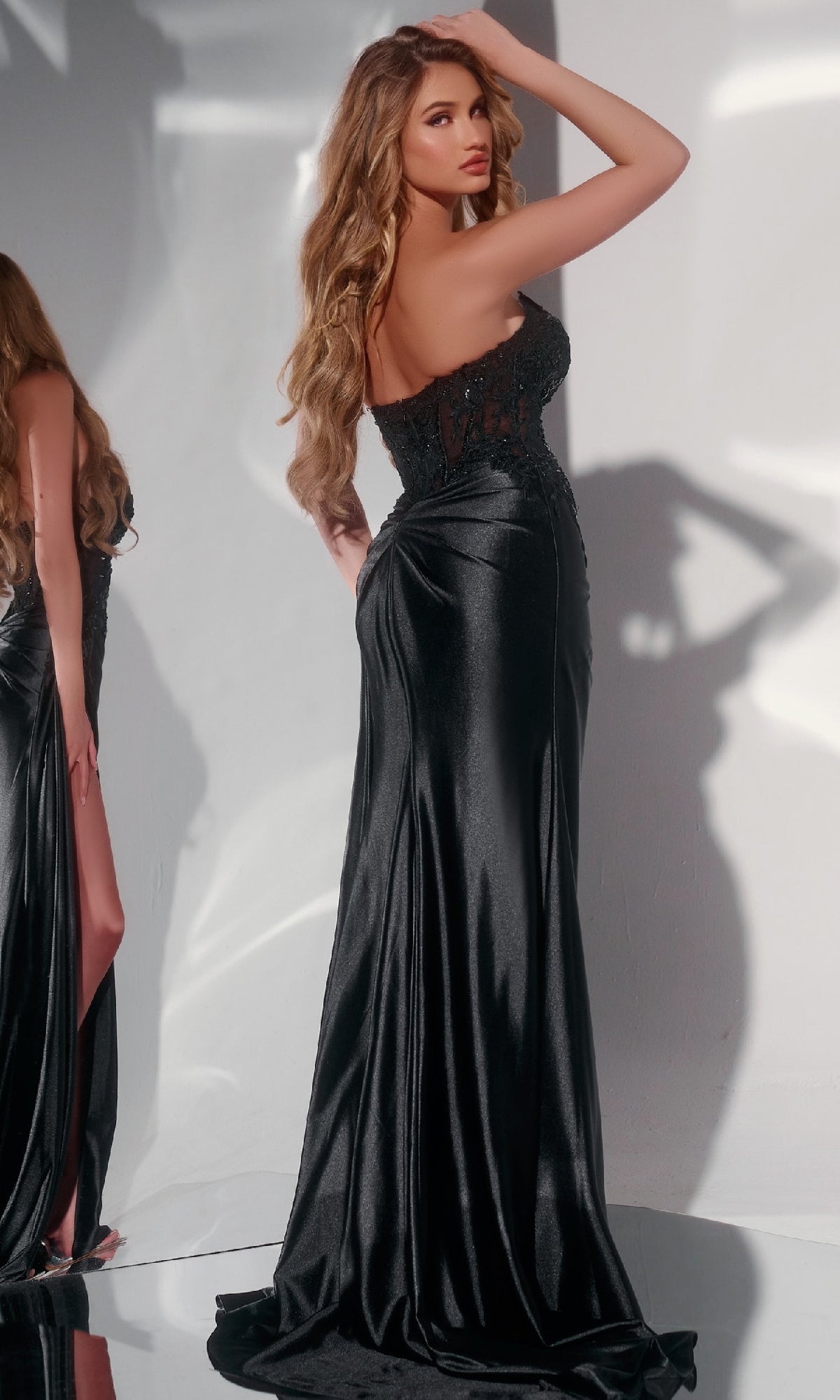 Wearing the Jovani 43848 Long Prom Dress, a woman poses with one hand on her head in front of a light gray wall. The strapless gown features a fitted bodice and flowing skirt.